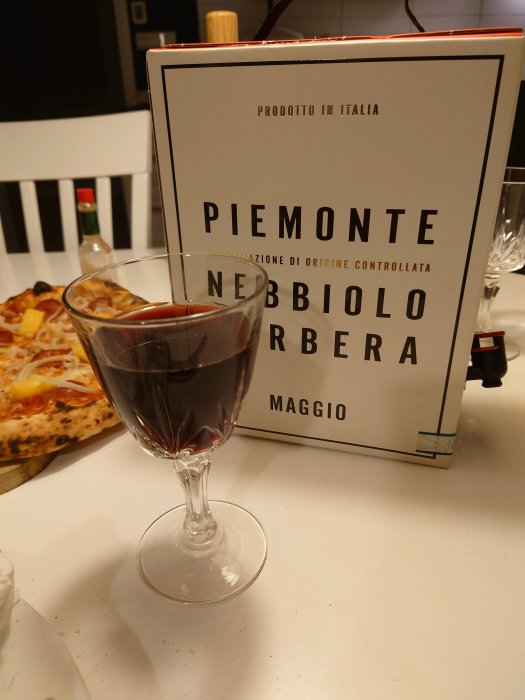 En italiensk Nebbiolo-vinbox från Piemonte bredvid ett glas rött vin, med en bit pizza i bakgrunden.