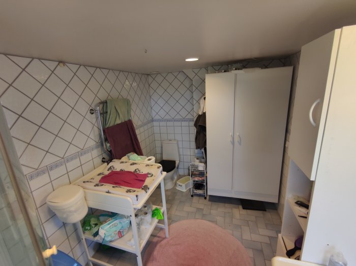 Interiör av ett badrum med vit kakel, möbler som skåp och skötbord, och en rosa matta på golvet.