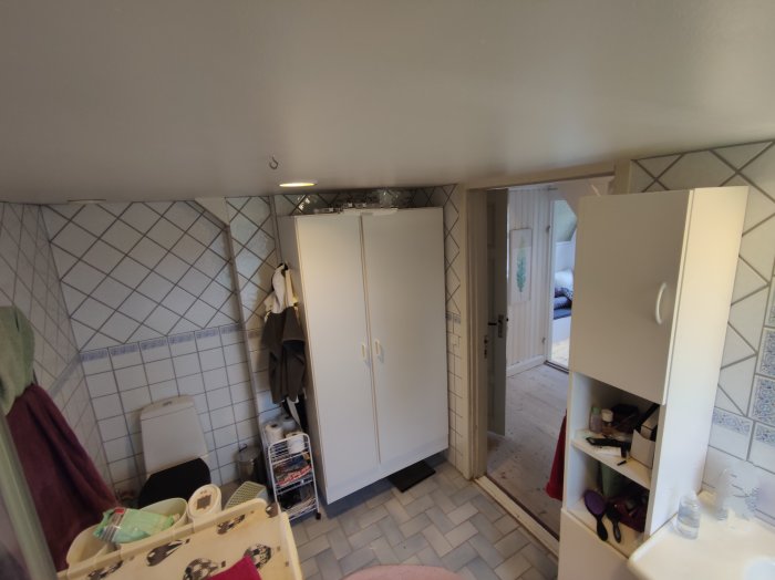 Ett badrum med vitt kakel, duschhörna och ett högskåp, dörr i bakgrunden leder till annat rum.