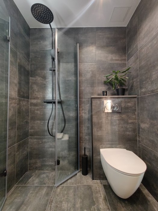 Nymodigt badrum med gråa kakelväggar, duschhörna, vägghängd toalett och provisoriska detaljer.