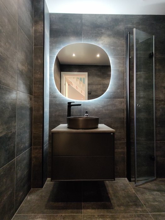 Modernt badrum med ett mörkt tvättställ och rund spegel, bredvid en dusch med glasdörrar.