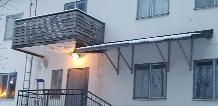 Tak över trappa med snö, intill balkong och husvägg med belysning.