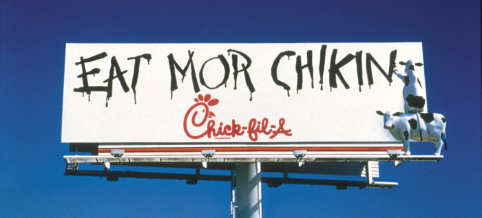 Reklamskylt med kor som målar "EAT MOR CHIKIN" på Chick-fil-A-logotypen mot klarblå himmel.