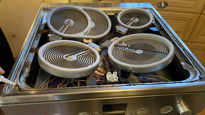 Öppnad elektrisk spis med borttagna kokplattor och synliga värmeslingor samt applicerat silikon på kanten.