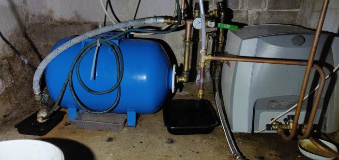 Blå hydrofor och vit varmvattenberedare i källarutrymme med synliga rör och ventiler.