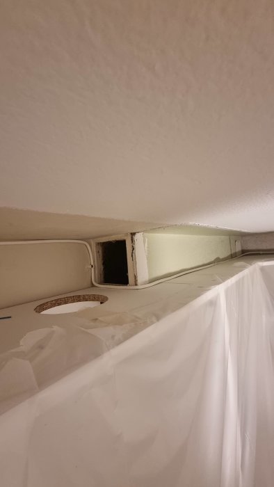 Vy under köksskåp där en kanalförlängning framför en befintlig frånluftsöppning syns, med plastskydd närmast.