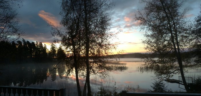 Soluppgång över en stilla sjö med ångande vatten, silhuetter av träd och färgglad himmel.