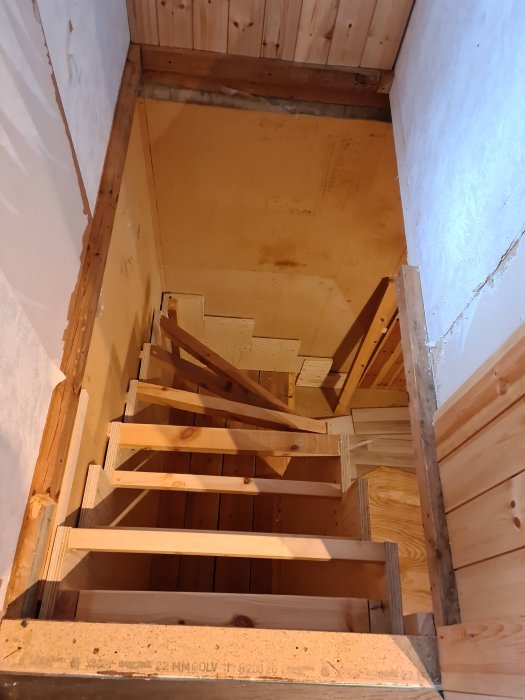 Nybyggd trätrappa sedd uppifrån utan sättsteg, i en ofärdig inomhusmiljö med träpanel och oavslutat nedre plan.