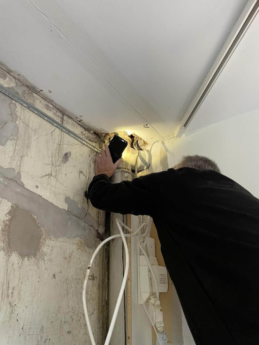 Person som inspekterar en öppnad vägg med synlig rörledning och elektriska kablar under renovering.