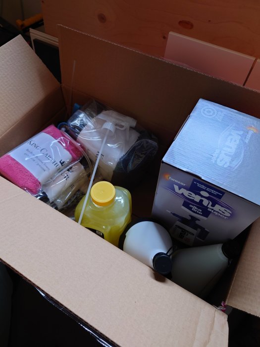 Öppen flyttkartong med diverse hushållsartiklar som rengöringsmedel, svampar och en Venus förpackning.