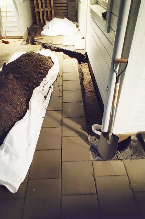 Grävd gräva längs husvägg för elinstallation i mörker, med spade och skyddsplast.