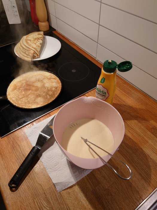 Stekning av pannkakor på spis med smet i skål och en flaska matolja på köksbänken.