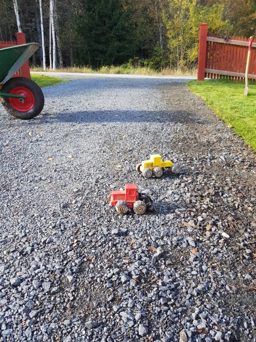 Infart med små leksakslastbilar på grus, gräskant och röd trädgårdsskjul i bakgrunden.