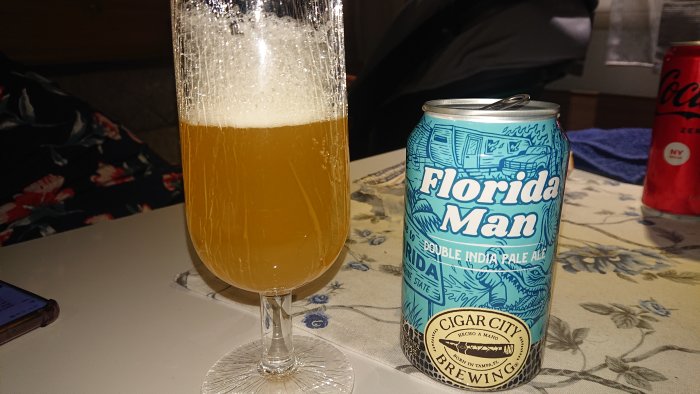 Ölglas med skum och ölburk "Florida Man Double IPA" på ett bord med mönstrad duk; Cola Zero-burk i bakgrunden.