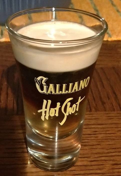 Ett glas Galliano Hot Shot med distinkta lager av kaffe och grädde på ett träbord.