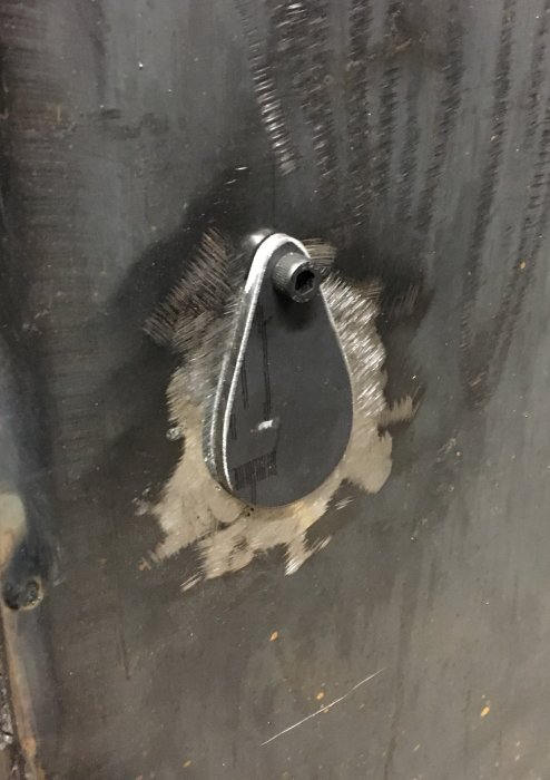 Metallplåt med spåren efter svetsning kring ett svart spjäll och en utstickande bult.