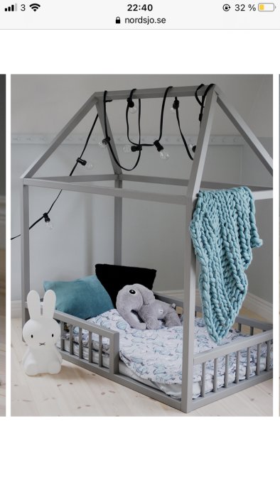 Barnsäng utformad som ett gråmålat hus med ljusslinga, mjukdjur, filtar och ett leksaksdjur framför sängen.