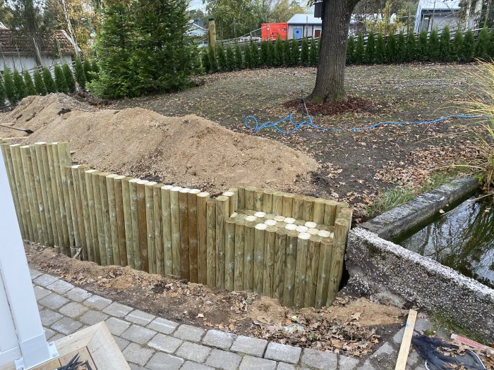 En nybyggd stödmur av vertikala trästolpar med en hög jordhög bakom i en trädgård.