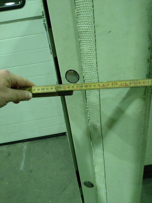 Hand håller ett måttband mot en vit garageport för att mäta portens tjocklek.