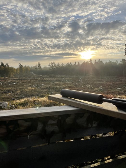 Soluppgång över ett öppet landskap med skog i bakgrunden, vy från en trädäck med byggnadsverktyg.