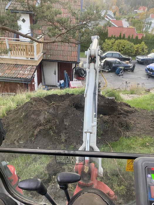 Vy från grävmaskinens hytt över en grävd grop nära ett hus, med grävarmens kontroller i förgrunden.