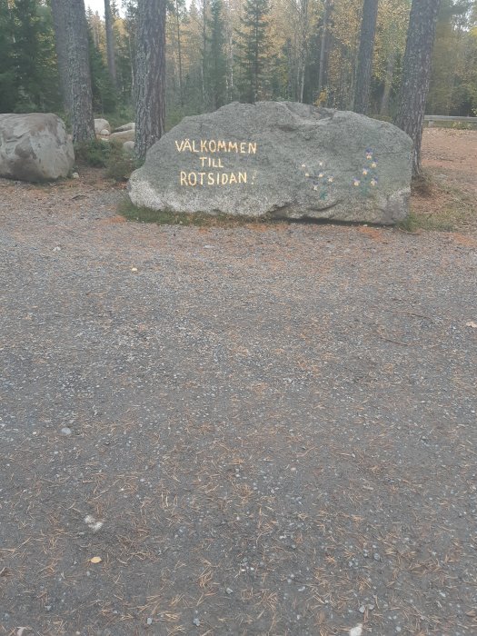 Stor sten med texten "VÄLKOMMEN TILL ROTSIDAN" i skogsmiljö.