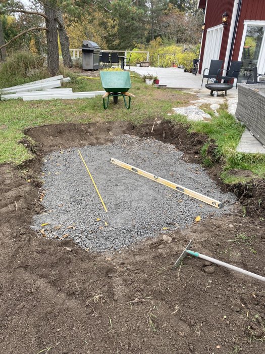 Grävd grop med grus för installation av spabad, trädgårdskärra och byggverktyg syns.