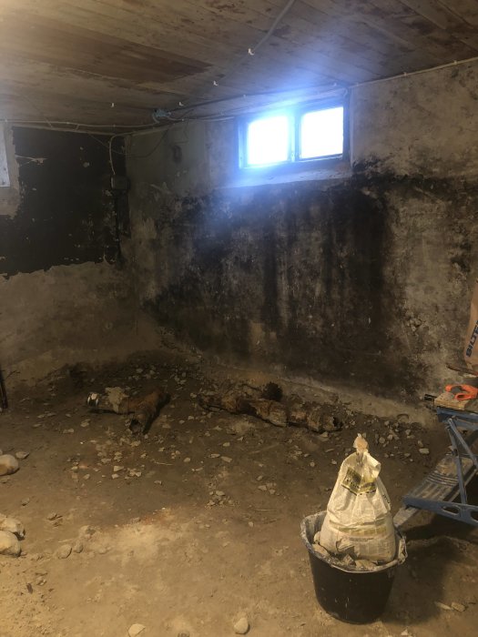 Basement under renovation with rough, damaged walls, exposed ceiling, debris on the floor, a small window, and renovation materials in the foreground.
