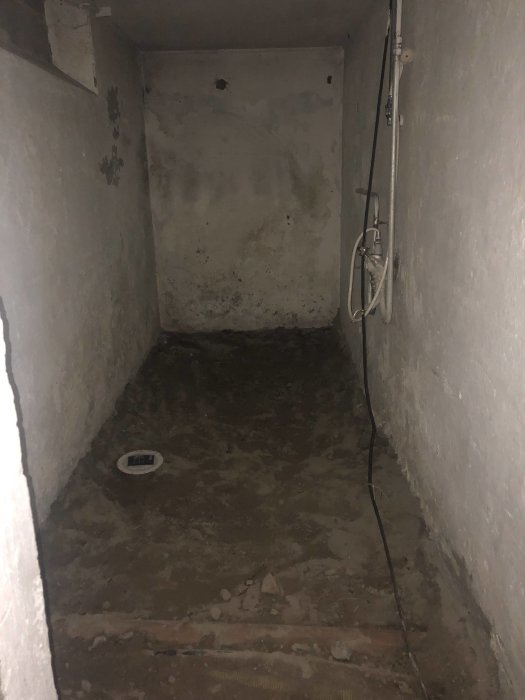 Basement under renovation with unfinished concrete floor and rough plastered walls. Visible are exposed pipes and a drainage hole.