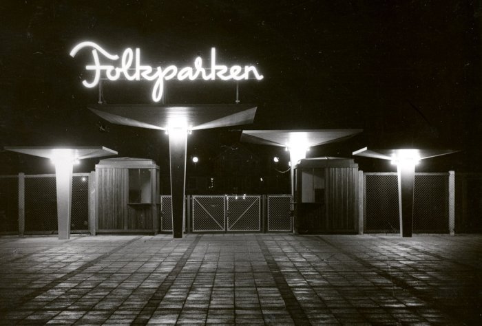 Svartvit bild av en tom entré till Folkets Park på natten med upplyst skylt som lyser "Folkparken".