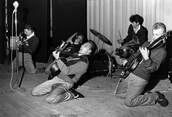 Svartvit bild av rockband som spelar, en man ligger ner och spelar gitarr medan andra musiker uppträder.