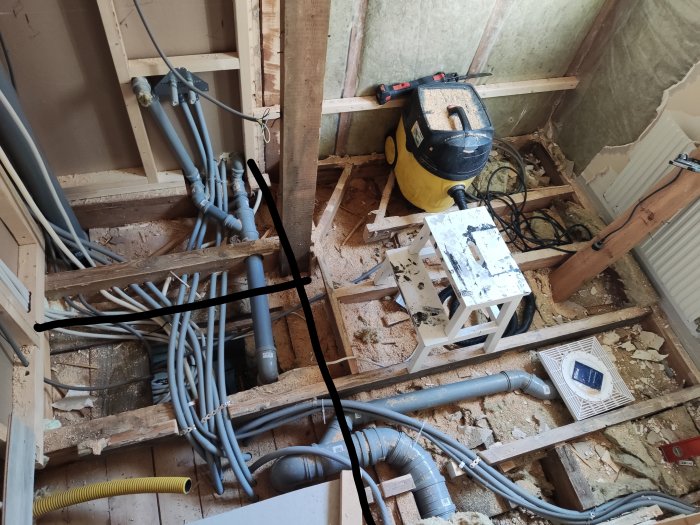 Rörigt byggprojekt med blottade bjälkar, rör och kablar på ett golv under renovering.