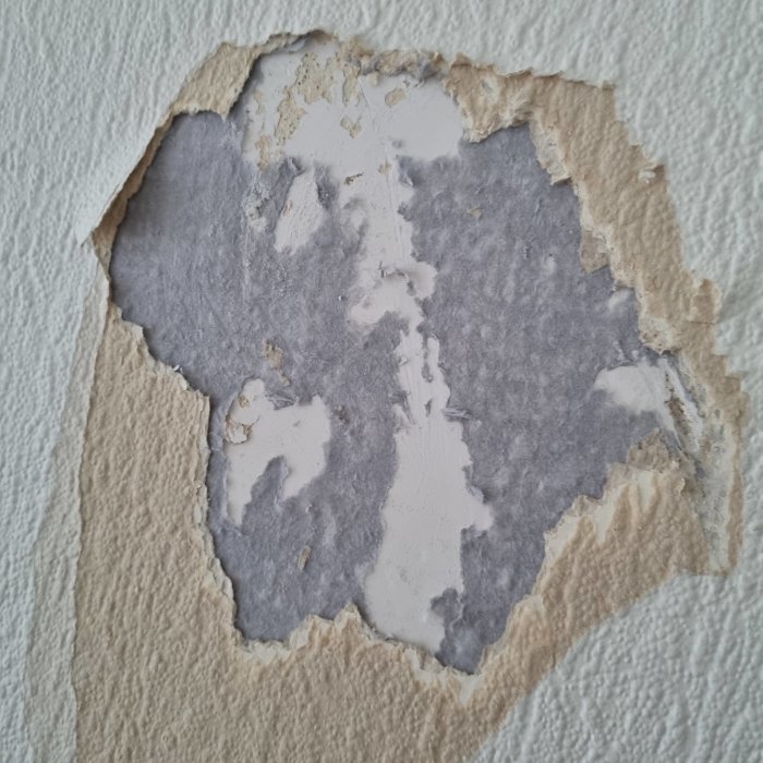 Rivet tapet avslöjar grått isoleringsliknande material på en vägg i ett vardagsrum.