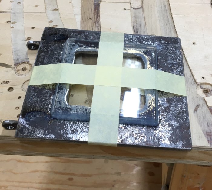 Grovmålad kaminlucka med genomskinligt glas fixerat med tejp på ett arbetsbord i en snickerilokal.