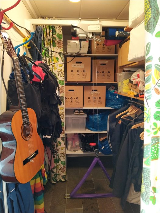 Rörigt förrådsutrymme med monterad vinsch, gitarr, kläder och diverse förvaringslådor.