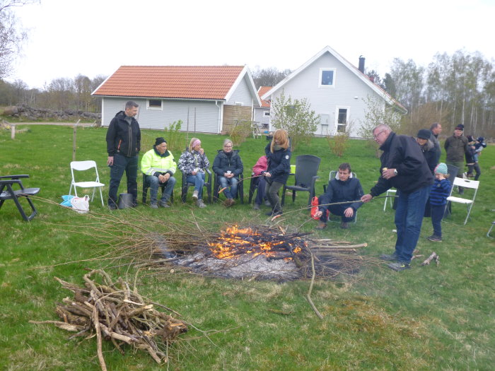 Människor samlas kring en brasa i en trädgård, hus med liggande panel i bakgrunden.