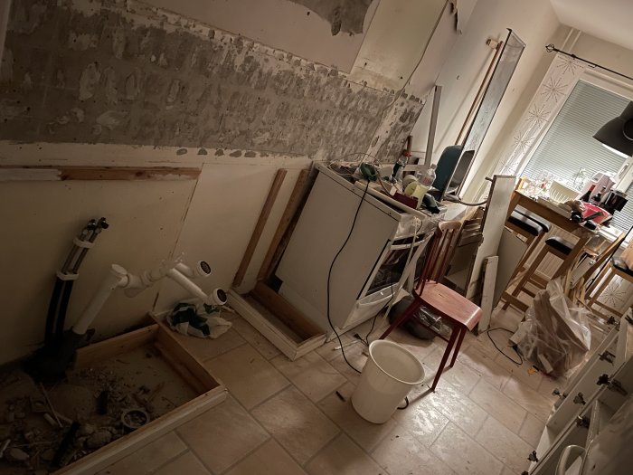 Kök under renovering med borttaget kakel och hyllor, spackel på vägg, och rörledningar synliga.