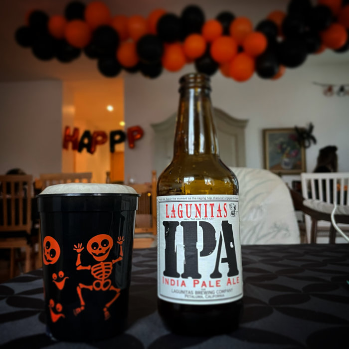 Flaska Lagunitas IPA bredvid pappmugg med skelettmönster, i rum med ballonger och "Happy"-dekoration i bakgrunden.