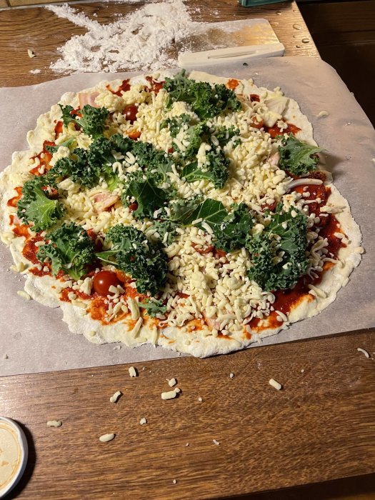 Glutenfri hemlagad pizza med tomat, ost, skinka och grönkål på bakplåtspapper med mjöl i bakgrunden.