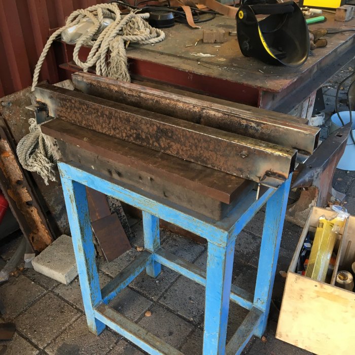 Rostiga metallbalkar monterade på en blå arbetsbock i ett rörigt verkstadsutrymme.