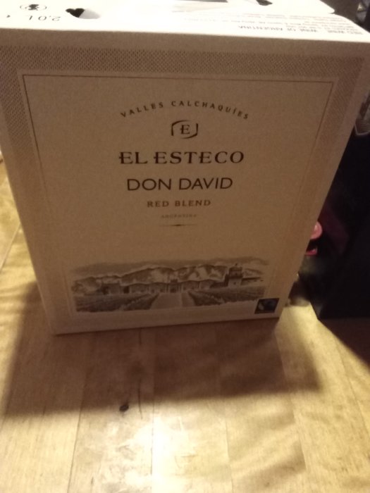 En kartong med rödvinet "El Esteco Don David Red Blend" står på ett trägolv.