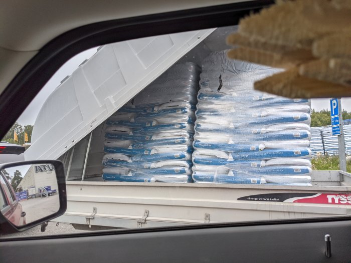 Vy från bilens förarsäte visar en annan bil överlastad med säckar av byggmaterial, osäkrade, sett genom ett öppet bakluckefönster.
