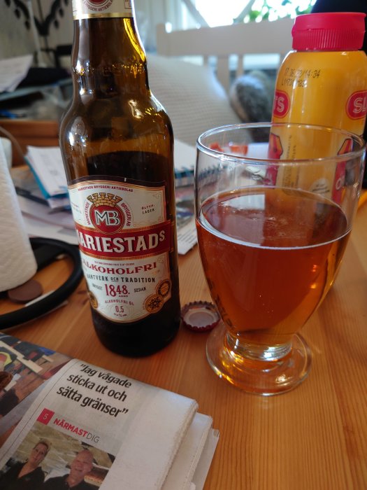 En flaska alkoholfri Mariestads öl bredvid ett glas med öl och en juiceflaska på ett bord med en tidning.