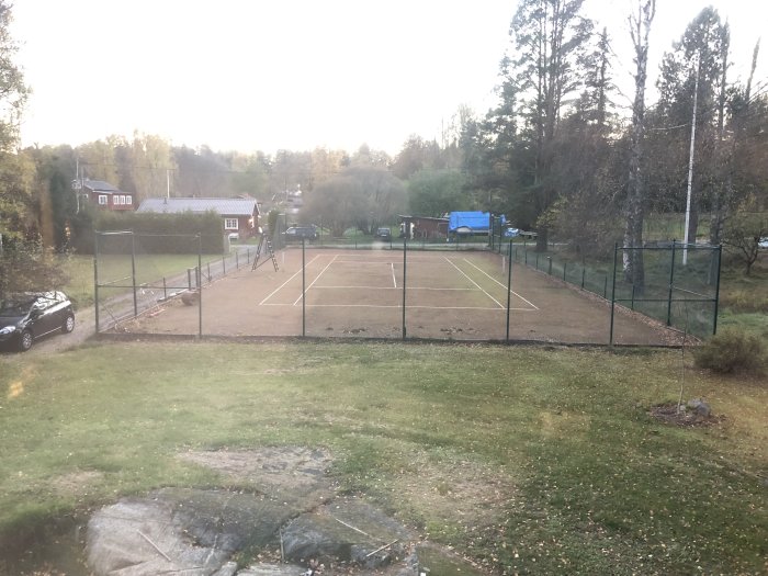 Tennisplan med stängsel, markerade linjer och omgivning som ska omvandlas till gräsmatta.