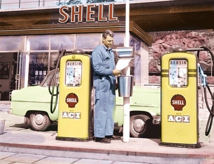 Man i arbetskläder vid Shellmack läser vid ACI-dekorerade bensinpumpar, vintagebil i bakgrunden.