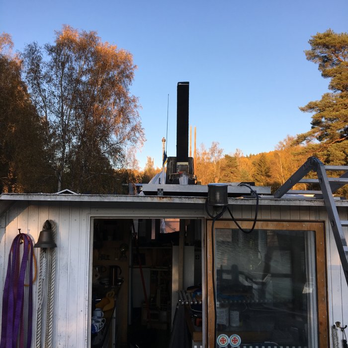 Skorsten genom taket på en verkstadsbyggnad omgiven av träd i solnedgång.