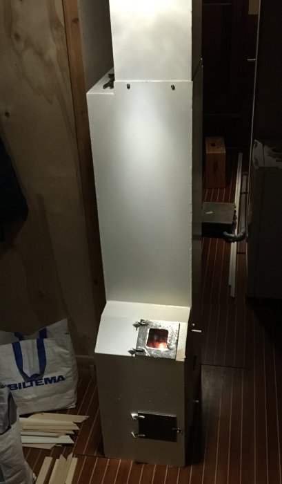 En metallkamin med en synlig eld genom ett litet fönster, installerad inomhus bredvid en sopsäck och byggmaterial.