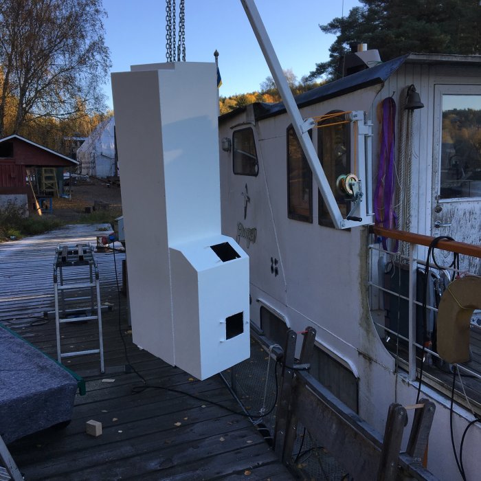 Vit skorsten som lyfts med kran på en båt, omgivet av byggredskap och skog i bakgrunden.