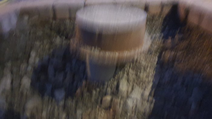Suddig bild av en brunn utgrävd nära hus, med ett grått rör, omgiven av jord och grus.