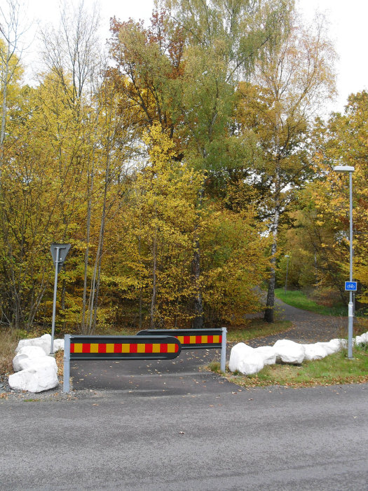 Bilspärr och trafikfålla vid en cykelväg omgiven av höstträd, för smal för breda cykelvagnar.